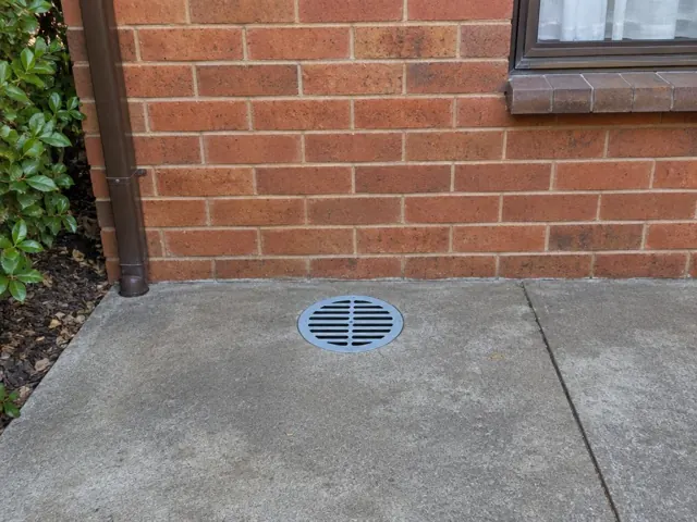 Round metal drain grate set in concrete beside a brick wall and window in Allambie Heights